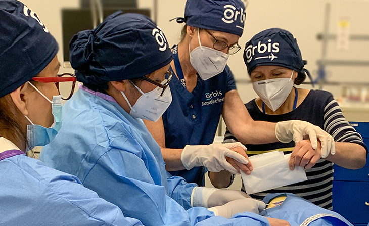 A woman wearing a blue surgical hat uses her hands to guide a woman putting a white drape on a manikin. 