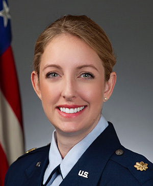 Blonde woman in Air Force uniform, light blue shirt, dark blue blazer and American flag in the background.