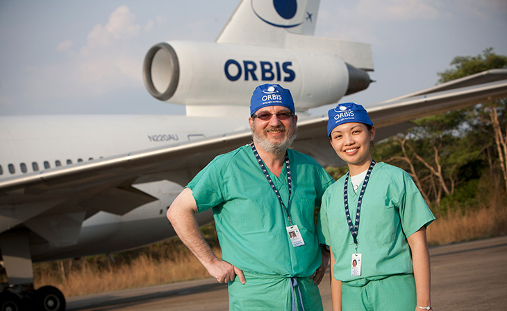 A man and woman wearing green medical scrubs stand in front of a white airplane with the word 
