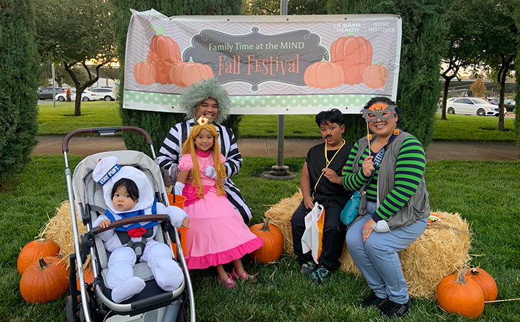 A family featuring two adults and three kids – one a baby in a stroller - pose on Hay Bales near a “Fall Festival” sign. They are dressed in Halloween costumes. 