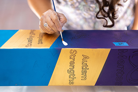 Close-up section of a colorful “Peace Pole” with the words “Autism Strengths” outlined and a woman’s hand starting to paint in white.