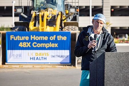 Man in scrubs and surgeon cap speaking on microphone at podium, sign in background states, “Future home of the 48X complex.”