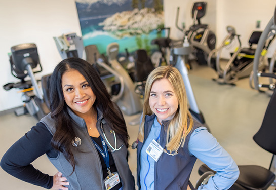 Veronica Encarnacion and Mallorie Mullendore wear blue scrubs and stand in front exercise machines