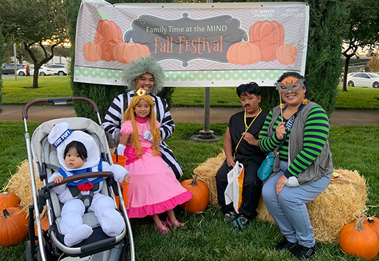 A family featuring two adults and three kids – one a baby in a stroller - pose on Hay Bales near a “Fall Festival” sign. They are dressed in Halloween