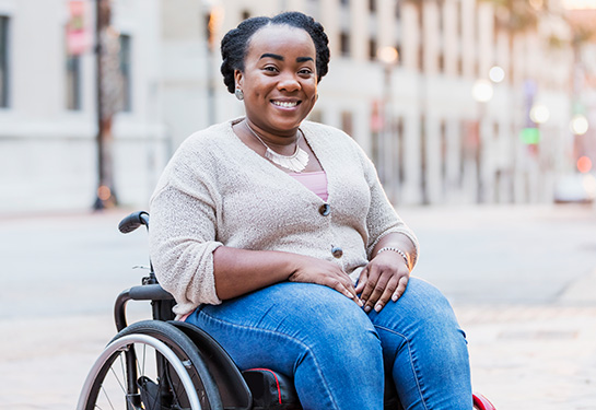 A mid adult Black woman in her 30s in a wheelchair outdoors, in the city, smiling at the camera. She has spina bifida.