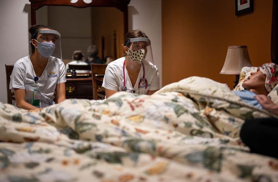 Two nursing students wearing masks, face shields and stethoscopes sit at the bedside of a mannikin simulation of a patient talking to their caregiver.
