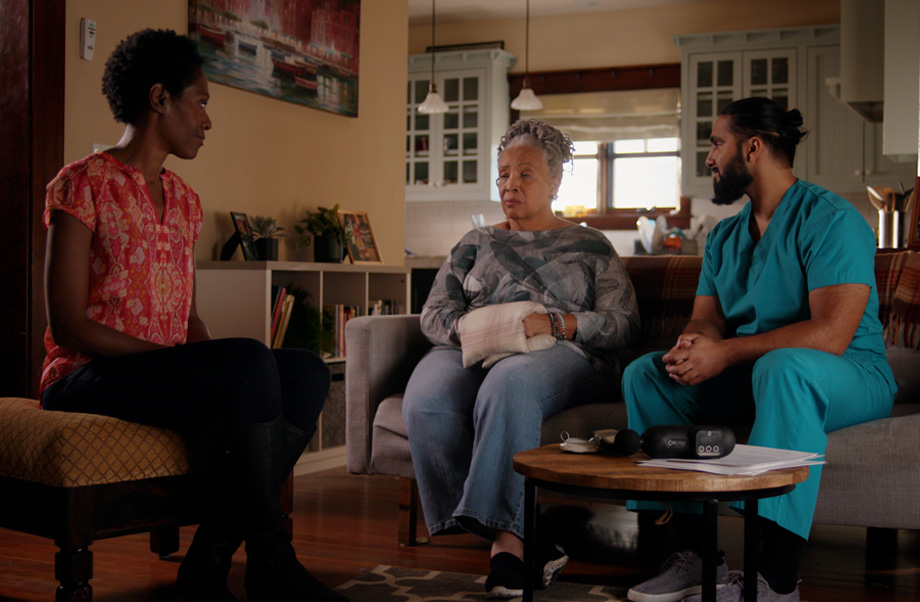 younger woman in red shirt on left sits on ottoman in living room talking to older woman in middle, who holds pillow in her hands, and man in scrubs on the left looking at older woman