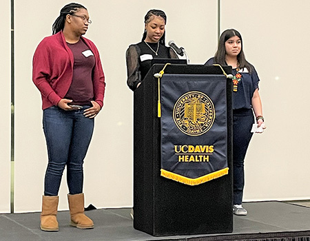 Three young women stand at a podium facing the audience