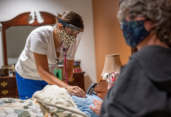 Nursing student wearing face shield and mask places stethoscope on the chest of a mannikin while family caregiver sits beside in bed 