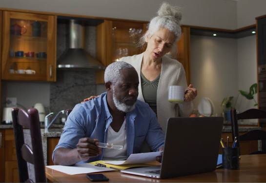 Older man looks at computer screen in front of him on kitchen island while older woman stands behind him looking at screen over his shoulder