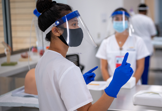 Nursing student wearing mask and faces hield looks right and points gloved finger while other nursing student in background works on manikin