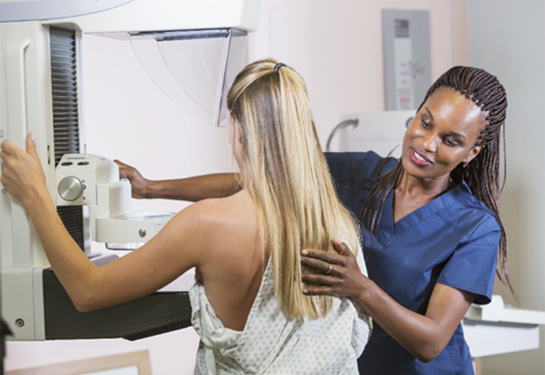A mature African American woman in her 40s wearing a hospital gown, getting her mammogram being helped by a technologist, a blond woman wearing scrubs