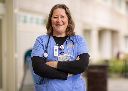 Chelsie Gilbeau wearing blue scrubs smiling and standing front of hospital with arms crossed