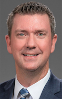 A smiling man with short, tidy brown hair wearing a blue business suit and a white collared shirt and a blue plaid tie