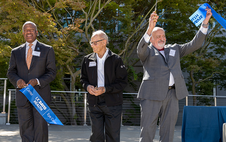 Three men, two in suits and ties and one wearing a dark zippered jacket, are holding scissors and pieces of a blue ribbon with the UC Davis Health Logo in white.