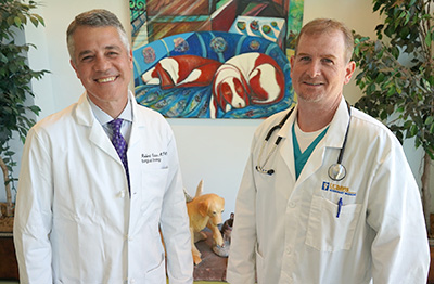 Robert J. Canter on the left and Dr. Robert B. Rebhun on the right, in front of a painting of two dogs lying on a couch next to two sculptures on a table, one of a yellow lab and one of a black chihuahua.