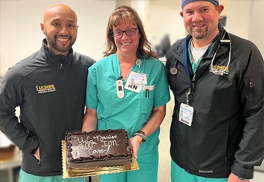 Two men and a woman wearing scrubs hold a brown cake with white writing that states, 'UC Davis 100th Ion Case!'