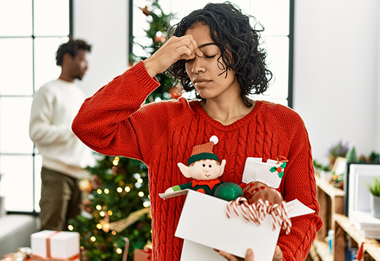 A tired looking woman wearing a red holiday sweater rubs the bridge of her nose.