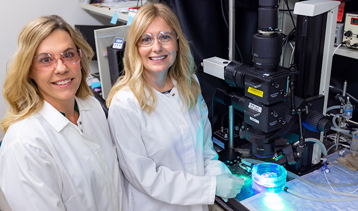 Two women with blond hair and in white lab coats and safety glasses stand in a lab next to a black imaging device with a glass dish underneath illuminated by blue and white lights. 