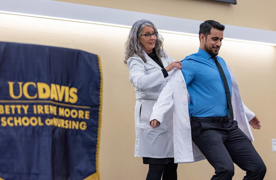 faculty member stands behind student and assists with the wearing of a white coat