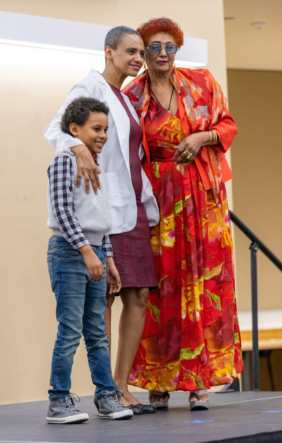 student in white coat stands in center posing on stage with right arm around son and left arm around mother