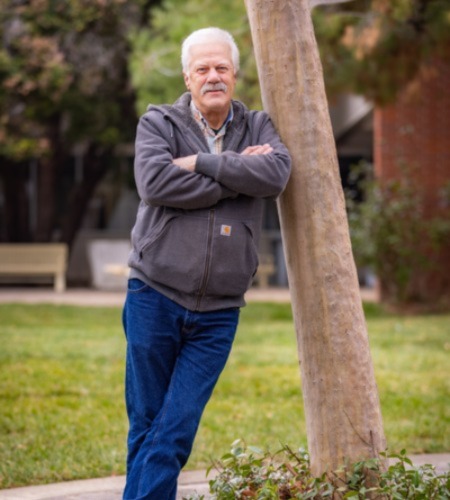 : A middle-aged man with white hair and a whitish-gray mustache, wearing a gray hooded sweatshirt and blue jeans leans against a tree trunk. 