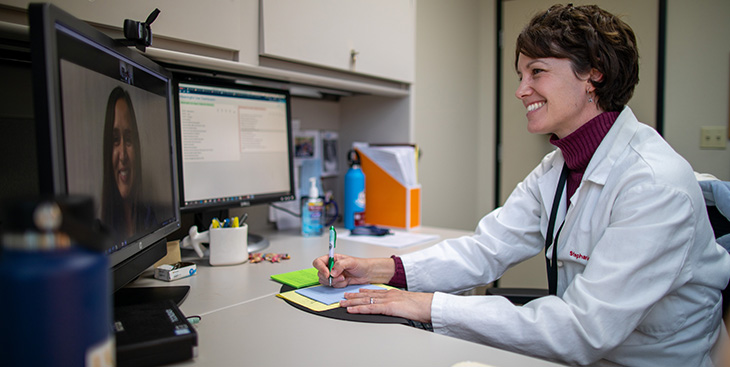 A white female doctor wearing a white coat taking notes as she looks at a camera and monitor during a telehealth session with a patient