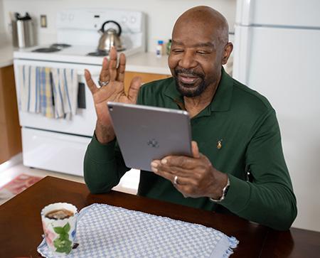 An African American man sitting at his kitchen table waving at a tablet. A cup of tea is in front of him.