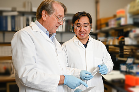 An older man with gray hair and a younger man with dark hair wearing white coats stand near each other as they work in a lab. 