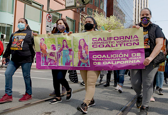 Four people wearing masks are walking down a city street and holding a yellow and purple sign with the words “California Domestic Workers Coalition.” 
