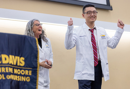 student wearing white coat raises arms and smiles while instructor, left, smiles