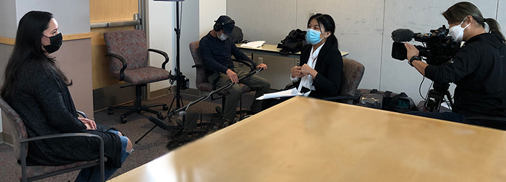 Woman with black mask sitting in chair looking into camera and being interviewed