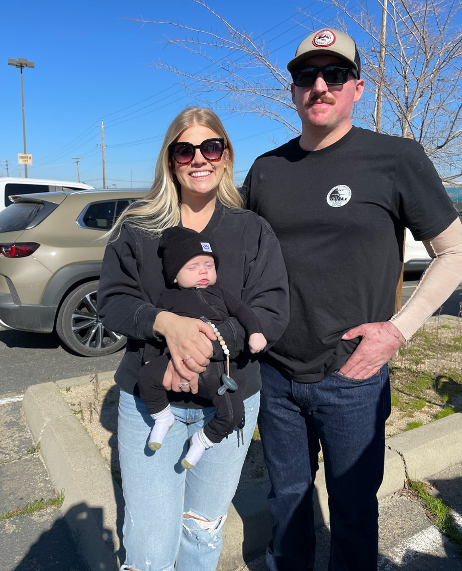 Woman, left, holds newborn baby in her arms, smiling, as husband stands beside her wearing a ballcap