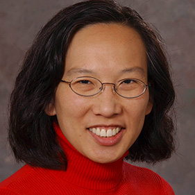 Woman with dark shoulder length hair, red turtleneck and glasses on gray background.
