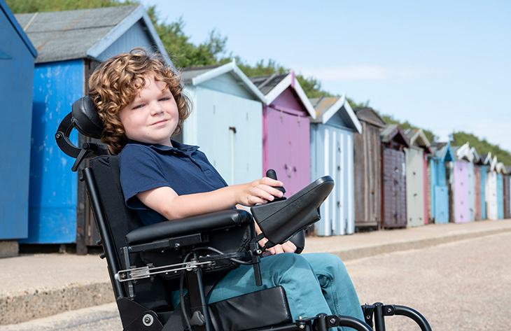 A six year old boy sitting in wheelchair in front of colorful painted beach huts, looking at camera, smiling