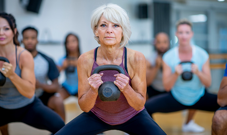 An older white woman doing a squat with a kettlebell, while participating in a co-ed, multi-ethnic, fitness class.