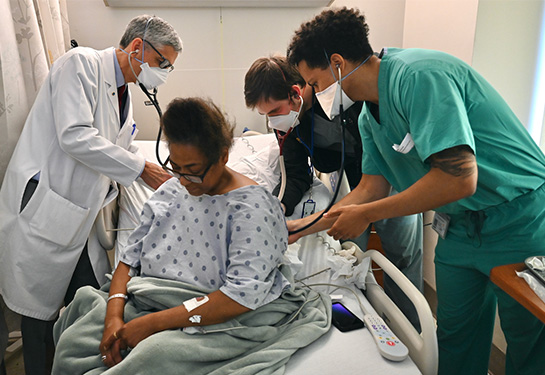 A doctor in white lab coat and two medical trainees use stethoscopes to check the lungs of a woman sitting on a hospital bed 