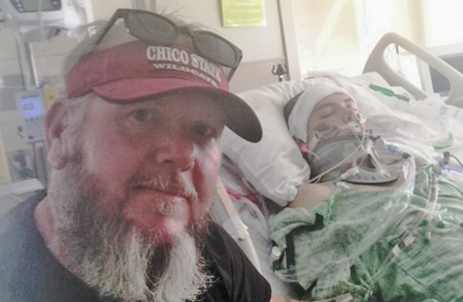 Bearded man with sunglasses and visor on top of his head sits next to hospital bed where he son lies in hospital gown and ventilator