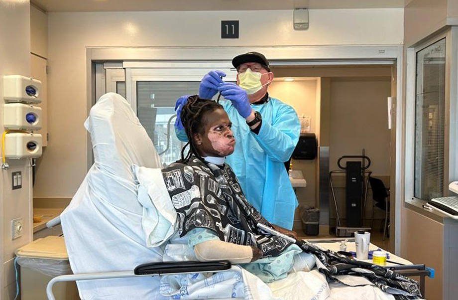 Man wearing mask and hospital gown stands next to and repairs a dreadlock on a recovering burn patient sitting in a ch