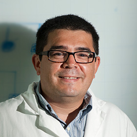Man with short dark hair stand before a white board wearing a lab coat and glasses. 