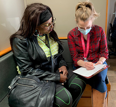 African American woman with headband and long, dark hair wearing a black outfit talking to blonde woman in plaid shirt who is filling out forms.