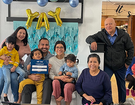 A man with short dark hair sits, surrounded by four other adults and three young children, including his daughter, holding a sign that reads “I matched UC Davis anesthesiology”