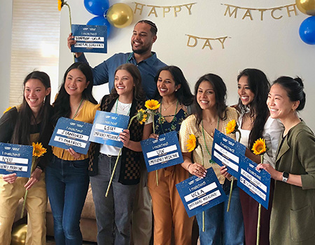 Eight medical students stand inside a home with blue signs announcing the residency programs they’ve been accepted to.