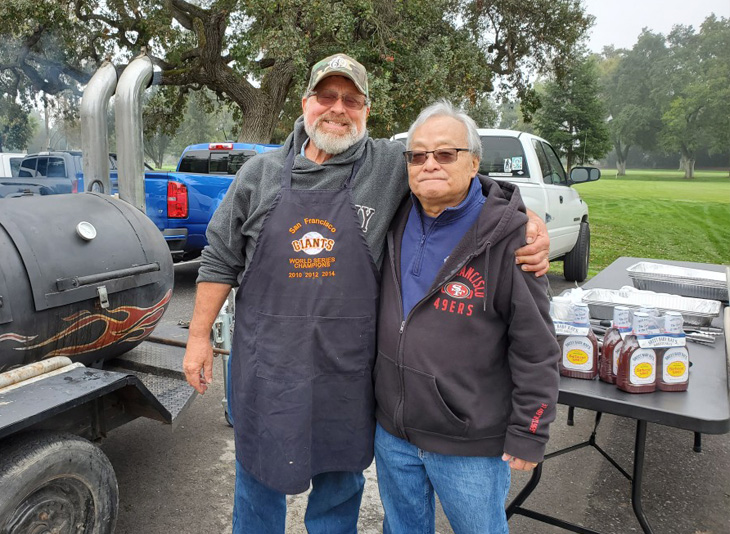 Two older men stand with their arms around each other’s shoulders in front of a barbecue pit