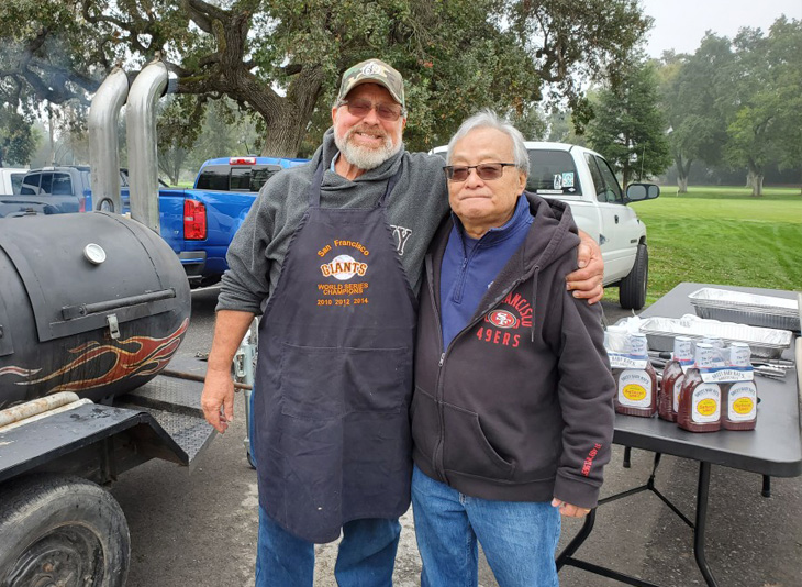 Two older men stand with their arms around each other's shoulders in front of a barbecue pit