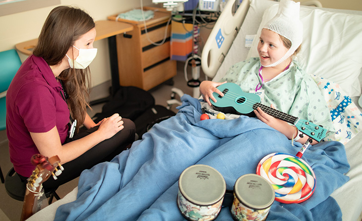 Woman sits by the bedside of a girl with a bandage on her head playing a ukulele in her hospital bed.