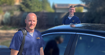 Home Infusion nurses Mark Brooks and Paul Harreld are dressed in navy blue scrubs and smiling while posing for a photo outside their vehicle near a patient's home. 