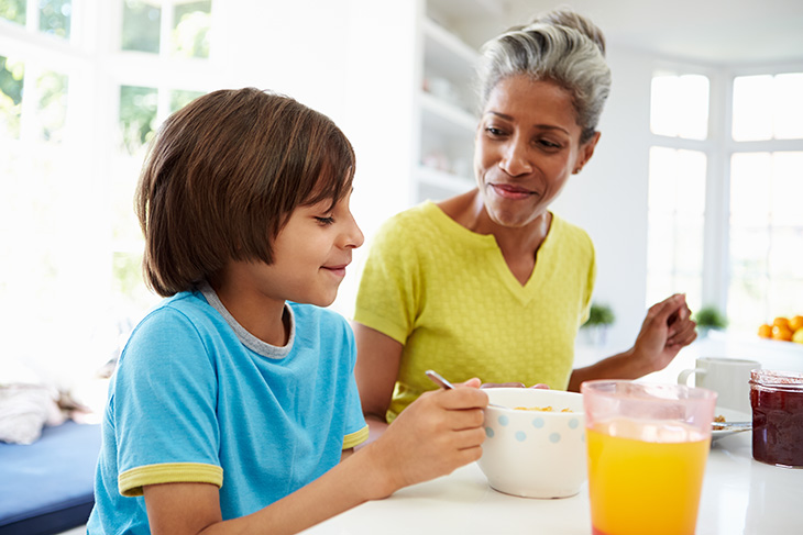 A grandmother smiles down at her grandson as they sit at a table eating breakfast together. The young boy eats from a large white bowl. A jar of jelly, glass of orange juice, white mug and plate are also in the table