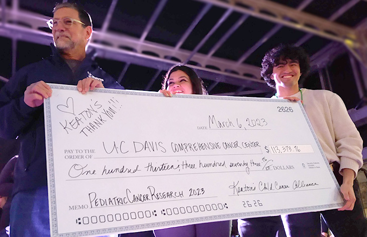 Three people on stage, man with dark hair accepts giant check from woman and young man.