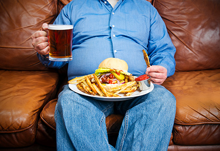 An overweight man sitting on a couch with a plate on his lap containing a hamburger and a pile of French fries. He holds a mug of beer in his hand. 